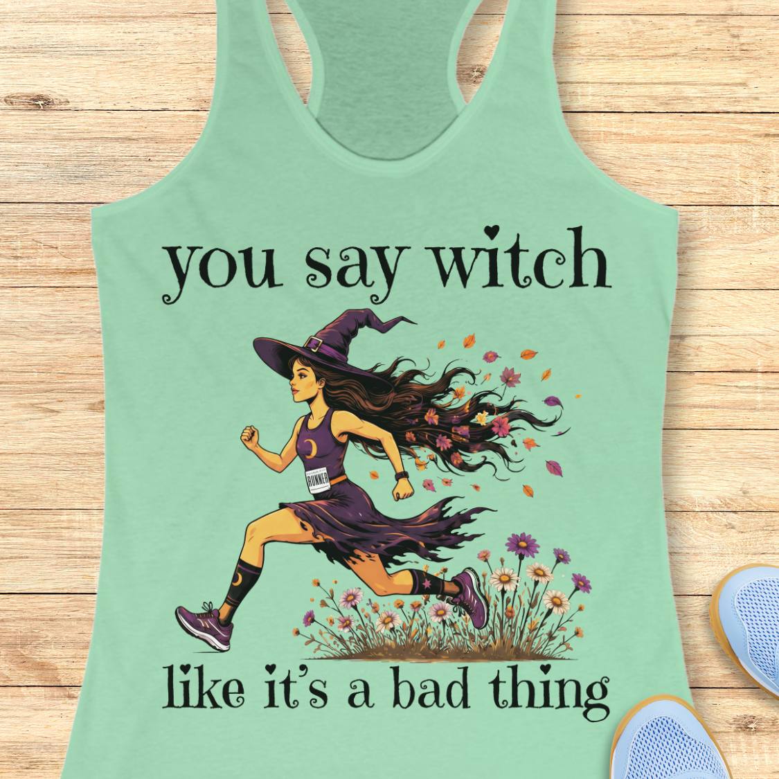 A mint green racerback tank top laid flat on a light wood background, featuring a graphic of a woman running as a witch with the text 'you say witch like it's a bad thing'.