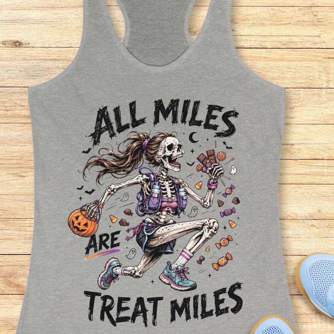 A grey racerback tank top laid flat on a light wood surface, featuring a colorful Halloween graphic of a skeleton girl holding a pumpkin and candy, with the text 'ALL MILES ARE TREAT MILES'. A partial blue shoe is visible at the bottom right.