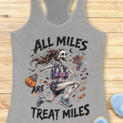 A grey racerback tank top laid flat on a light wood surface, featuring a colorful Halloween graphic of a skeleton girl holding a pumpkin and candy, with the text 'ALL MILES ARE TREAT MILES'. A partial blue shoe is visible at the bottom right.