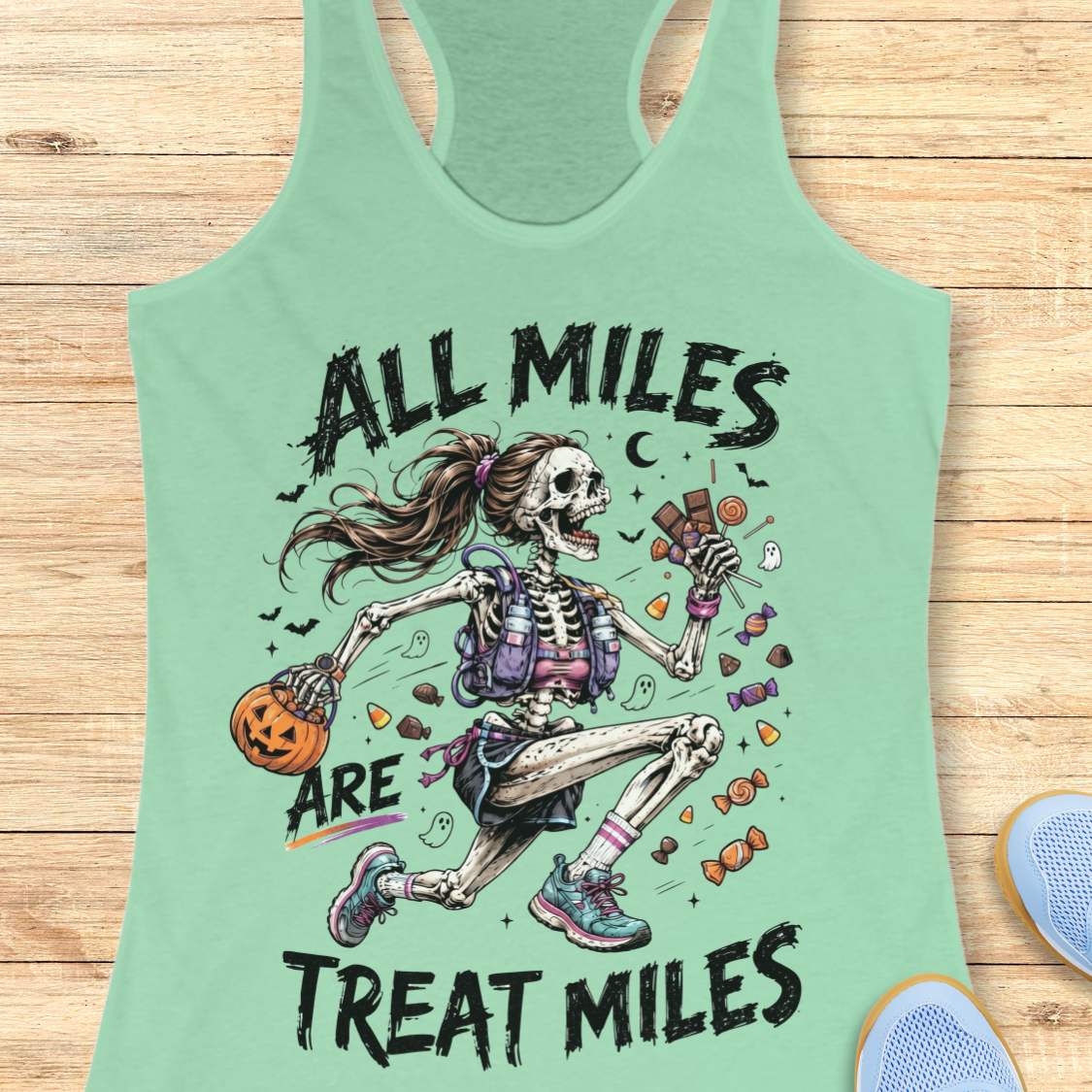 A mint green racerback tank top laid flat on a light wood background, featuring a colorful graphic of a running skeleton holding candy and a pumpkin, with the text 'ALL MILES ARE TREAT MILES'.