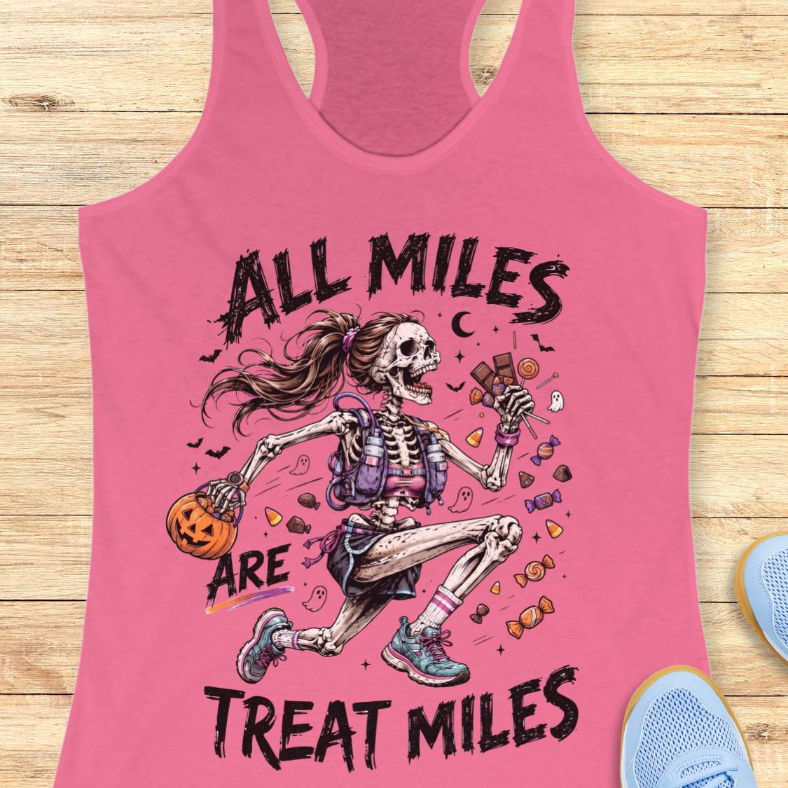 A pink racerback tank top laid flat on a light wood surface, featuring a colorful graphic of a running skeleton holding a pumpkin and candy, with the text 'ALL MILES ARE TREAT MILES.' Partial blue shoes are visible at the bottom right as styling props.