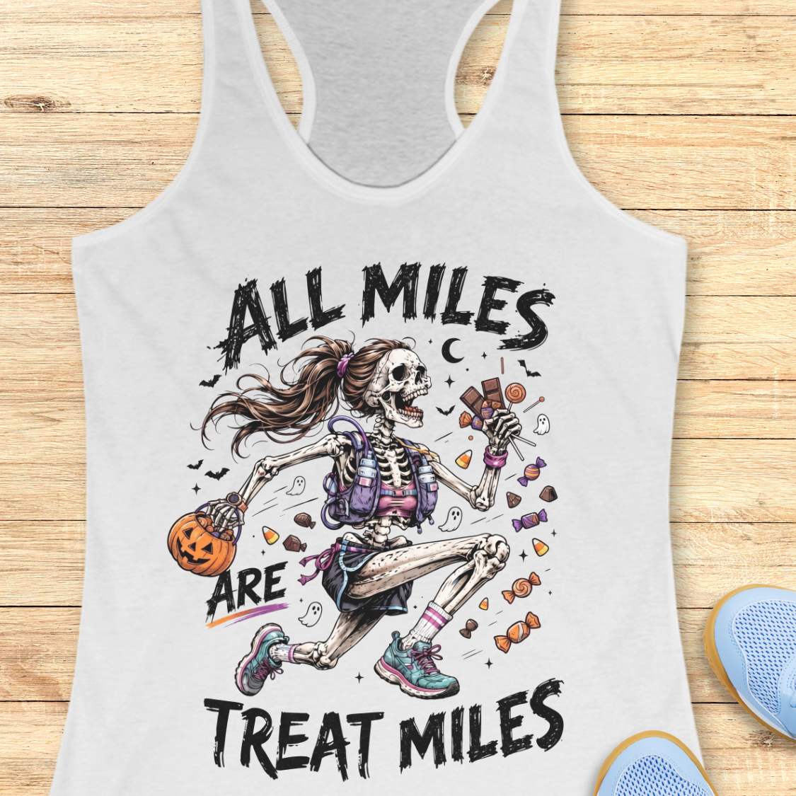 A white racerback tank top laid flat on a light wood surface, featuring a colorful Halloween graphic of a skeleton girl holding a pumpkin and candy, with the text 'ALL MILES ARE TREAT MILES'. A partial blue shoe is visible at the bottom right.