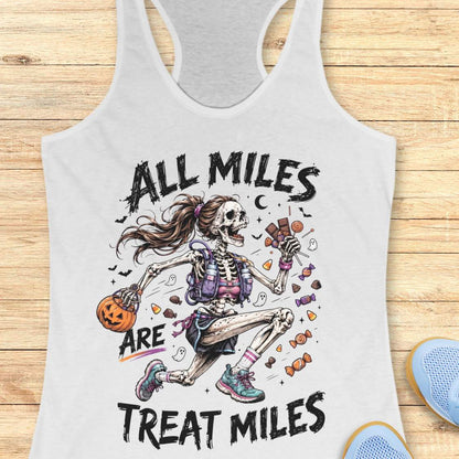 A white racerback tank top laid flat on a light wood surface, featuring a colorful Halloween graphic of a skeleton girl holding a pumpkin and candy, with the text 'ALL MILES ARE TREAT MILES'. A partial blue shoe is visible at the bottom right.