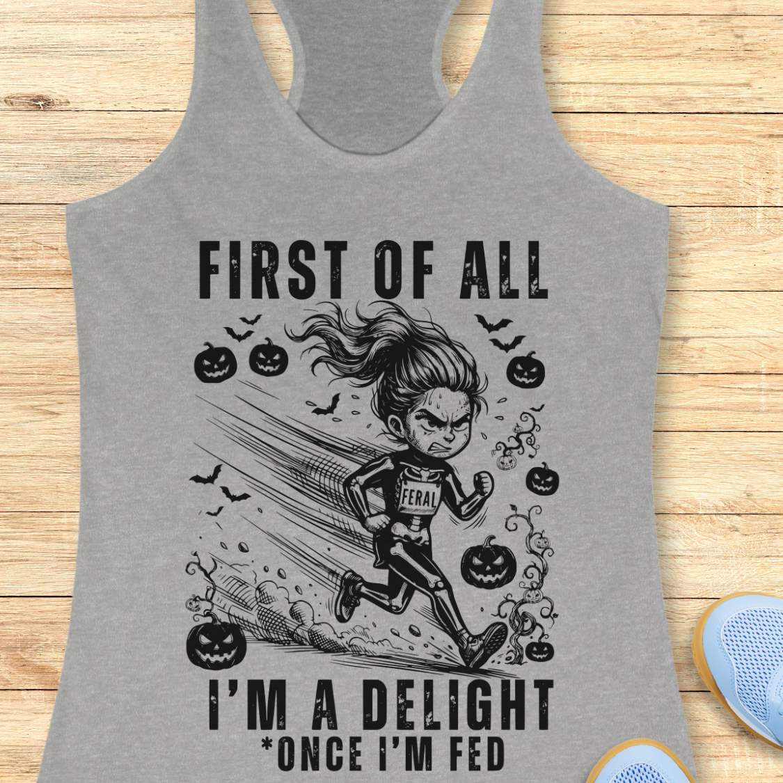 A heather gray racerback tank top with a black graphic print of a girl running while chasing jack-o'-lanterns and bats, with the text 'FIRST OF ALL I'M A DELIGHT *ONCE I'M FED', displayed flat on a light wood background.