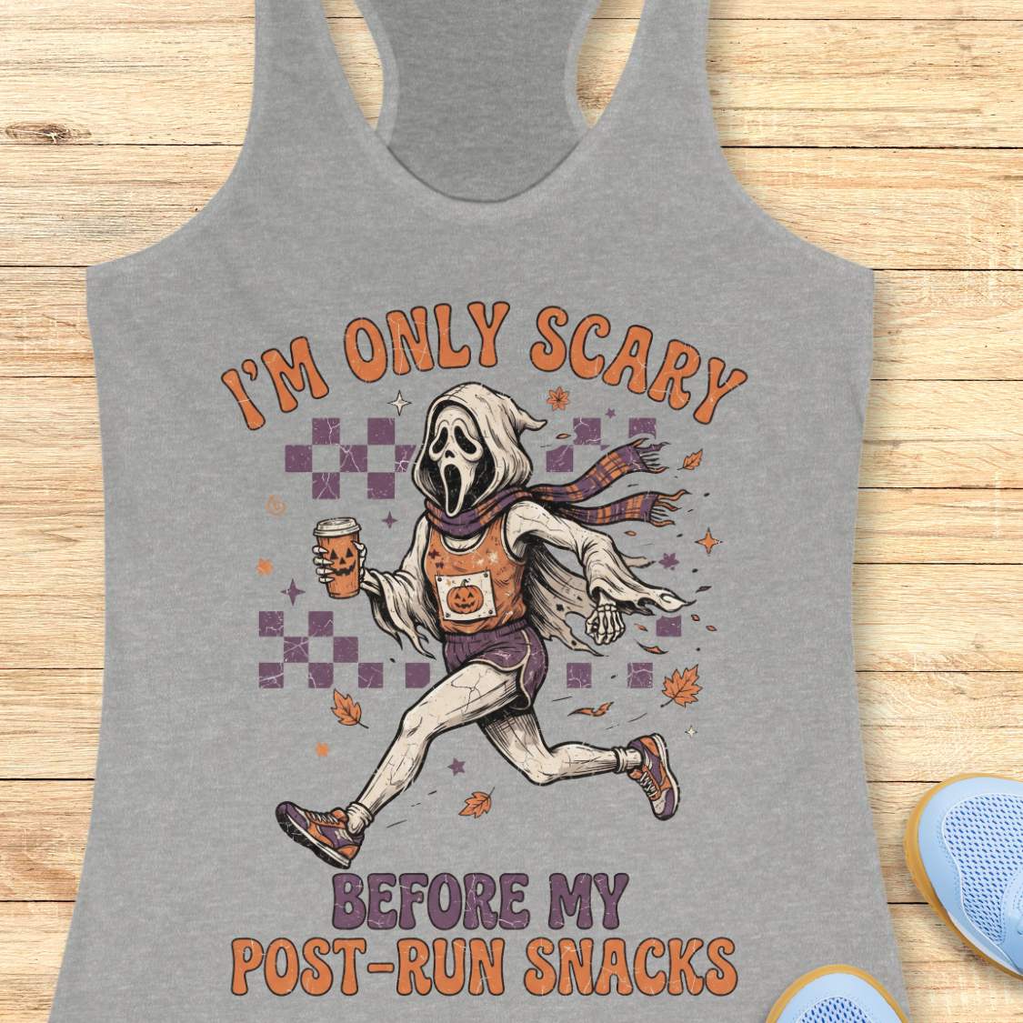 A heather gray racerback tank top laid flat on a light wood surface, featuring a Halloween-themed graphic of a ghostly figure running with a mug and the text 'I'M ONLY SCARY BEFORE MY POST-RUN SNACKS'.