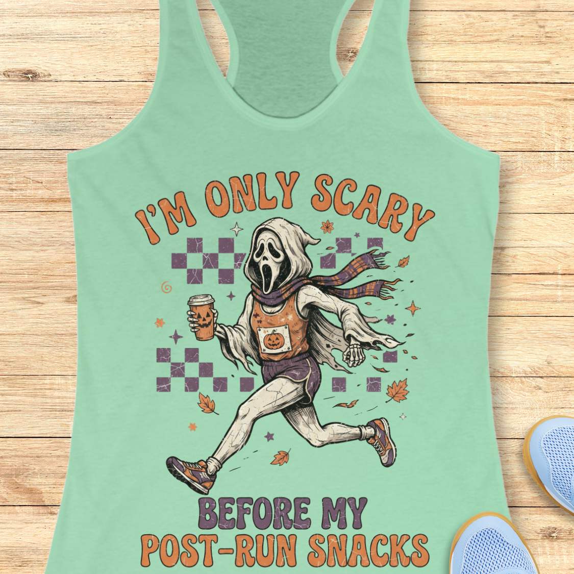 A mint green racerback tank top laid flat on a light wood background, featuring a graphic of a Ghostface-like running figure holding a coffee cup with the text 'I'M ONLY SCARY BEFORE MY POST-RUN SNACKS'.