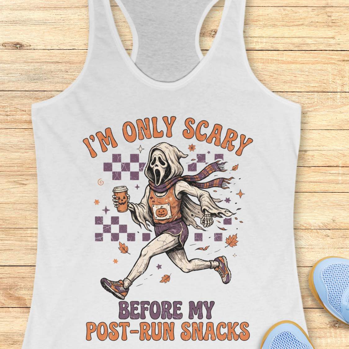 A white racerback tank top laid flat on a light wood background, featuring a graphic of Ghostface running with a snack and the text 'I'M ONLY SCARY BEFORE MY POST-RUN SNACKS'. Part of a blue shoe is visible in the bottom right corner.
