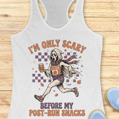 A white racerback tank top laid flat on a light wood background, featuring a graphic of Ghostface running with a snack and the text 'I'M ONLY SCARY BEFORE MY POST-RUN SNACKS'. Part of a blue shoe is visible in the bottom right corner.