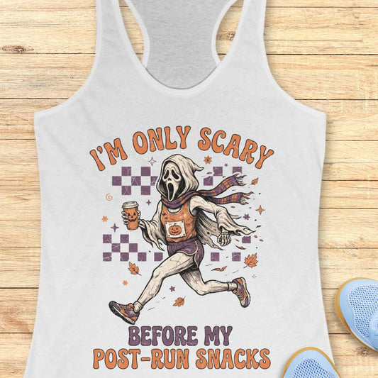 A white racerback tank top laid flat on a light wood background, featuring a graphic of Ghostface running with a snack and the text 'I'M ONLY SCARY BEFORE MY POST-RUN SNACKS'. Part of a blue shoe is visible in the bottom right corner.