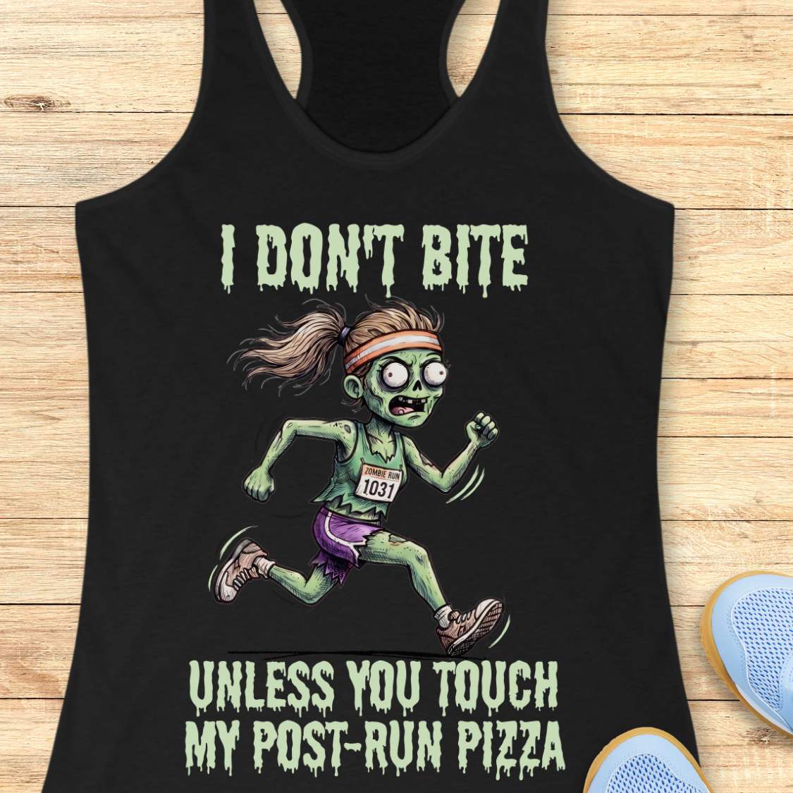A black racerback tank top laid flat on a light wood surface, featuring a zombie running graphic with the text 'I DON'T BITE UNLESS YOU TOUCH MY POST-RUN PIZZA.' Partial blue shoes appear at the bottom right as styling props.