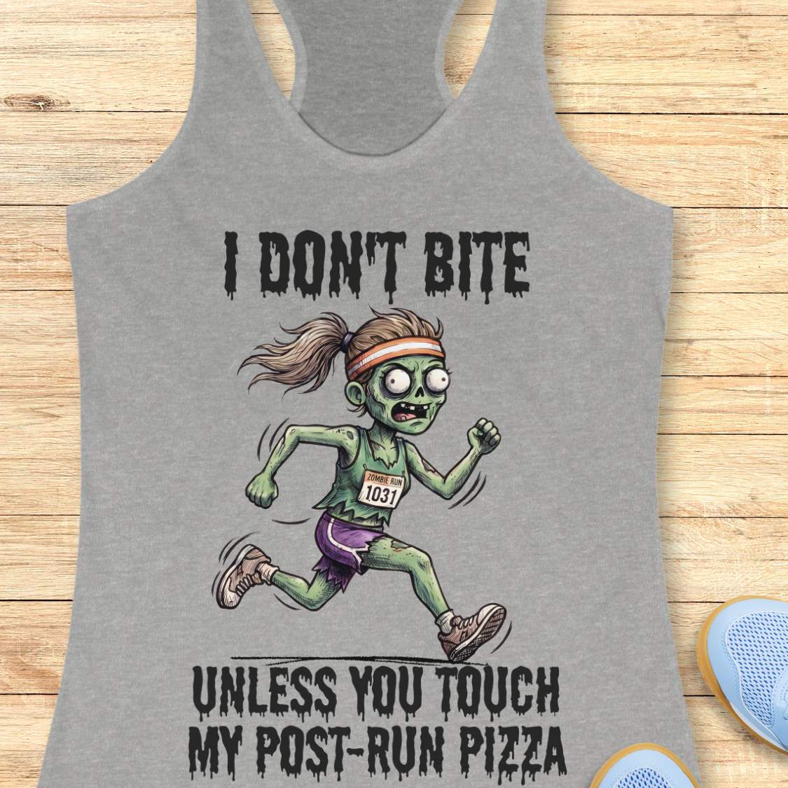 A heather gray racerback tank top laid flat on a light wood surface, featuring a zombie running graphic with the text 'I DON'T BITE UNLESS YOU TOUCH MY POST-RUN PIZZA.' A pair of blue sneakers is partially visible at the bottom right as a styling prop.