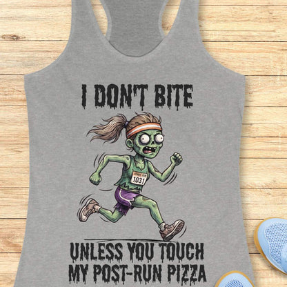 A heather gray racerback tank top laid flat on a light wood surface, featuring a zombie running graphic with the text 'I DON'T BITE UNLESS YOU TOUCH MY POST-RUN PIZZA.' A pair of blue sneakers is partially visible at the bottom right as a styling prop.