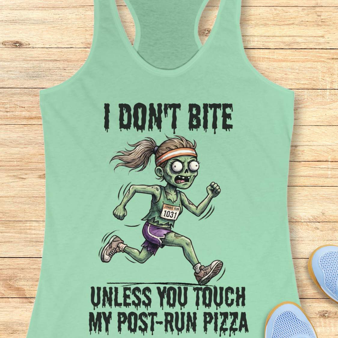A mint green racerback tank top laid flat on a light wood background, featuring a cartoon zombie runner graphic with the text 'I DON'T BITE UNLESS YOU TOUCH MY POST-RUN PIZZA.' A blue shoe is partially visible at the bottom right as a styling prop.