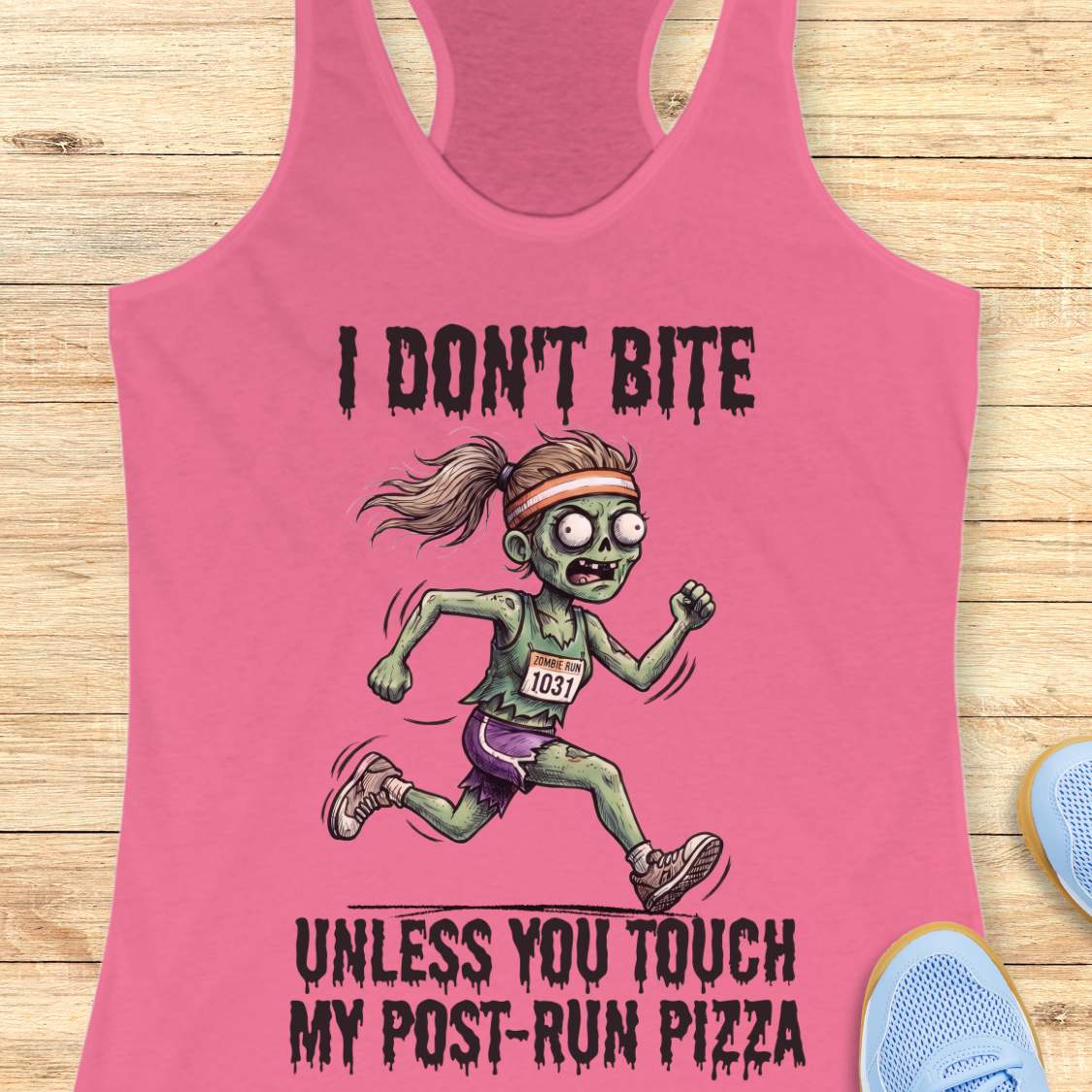 A pink racerback tank top laid flat on a light wood surface, featuring a cartoon zombie runner graphic with the text 'I DON'T BITE UNLESS YOU TOUCH MY POST-RUN PIZZA.' A pair of blue running shoes is partially visible at the lower right edge.