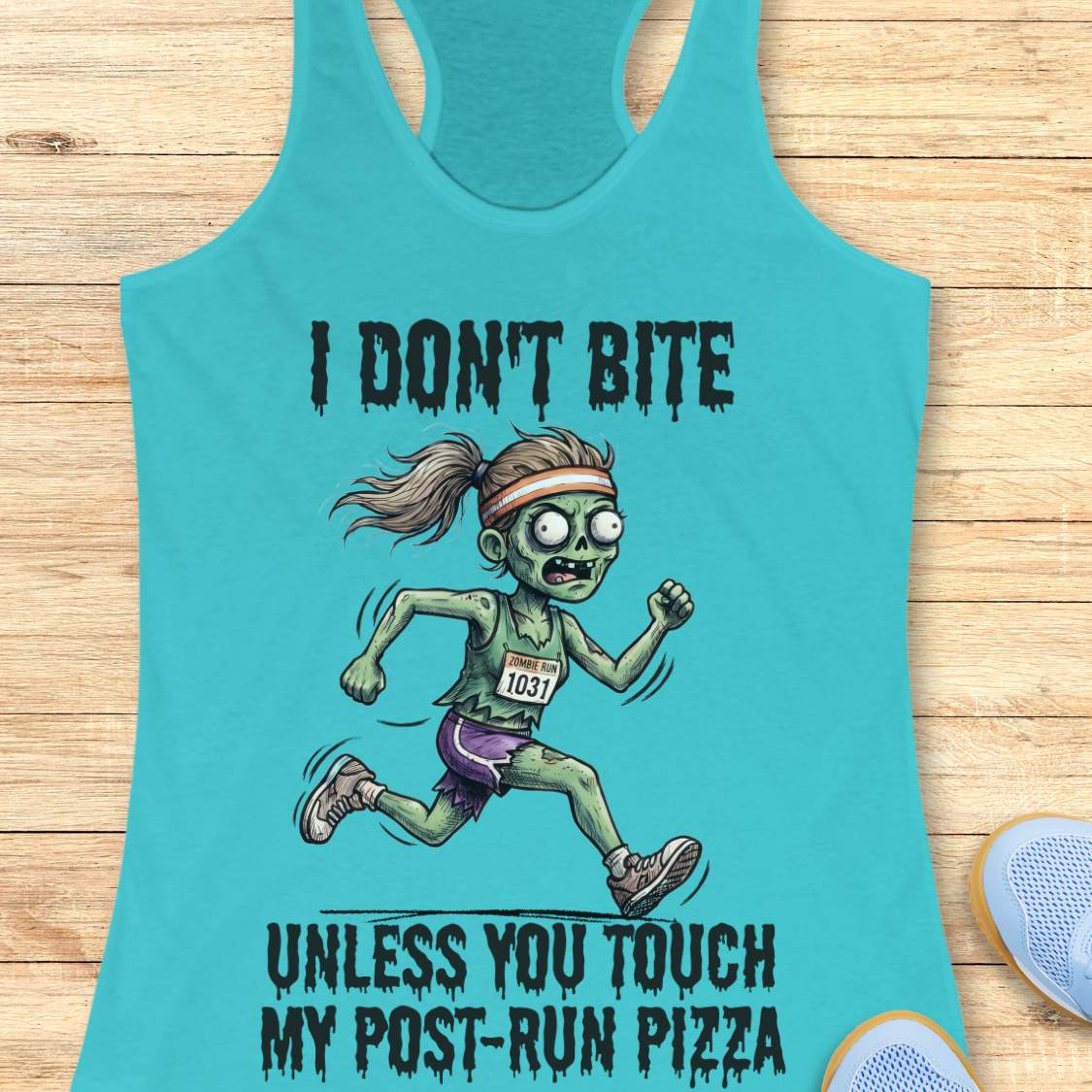 A turquoise racerback tank top laid flat on a light wood surface, featuring a zombie running graphic with the text 'I DON'T BITE UNLESS YOU TOUCH MY POST-RUN PIZZA.' Partial blue shoes are visible at the bottom right as styling props.