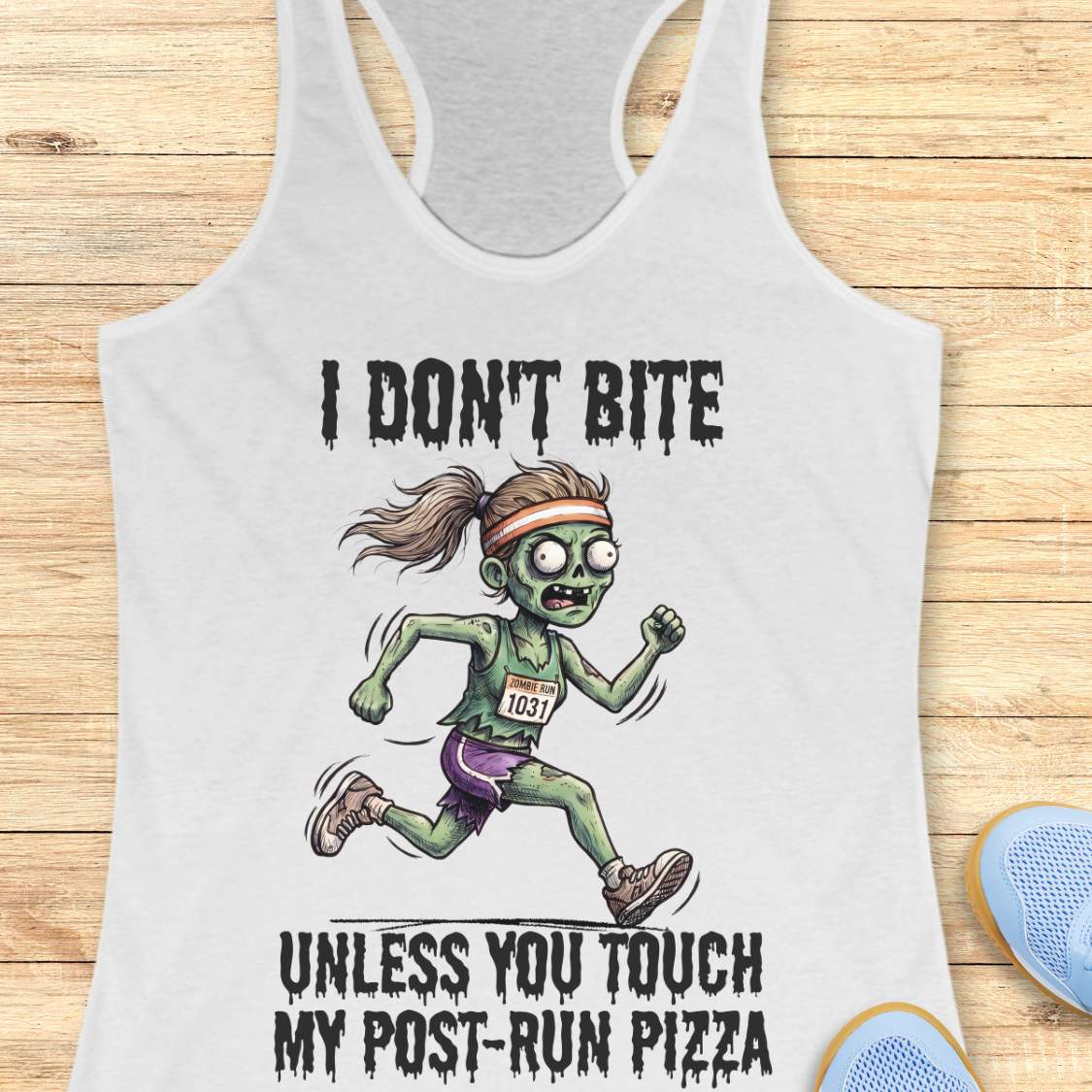 A white racerback tank top laid flat on a light wood background, featuring a zombie runner graphic with the text 'I DON'T BITE UNLESS YOU TOUCH MY POST-RUN PIZZA.' Partial blue shoes are visible at the bottom right as styling props.