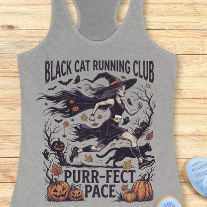 A heather gray racerback tank top laid flat on a light wood surface, featuring a Halloween graphic of a witch running with a black cat, pumpkins, bats, and the text 'BLACK CAT RUNNING CLUB' and 'PURR-PECT PACE'.