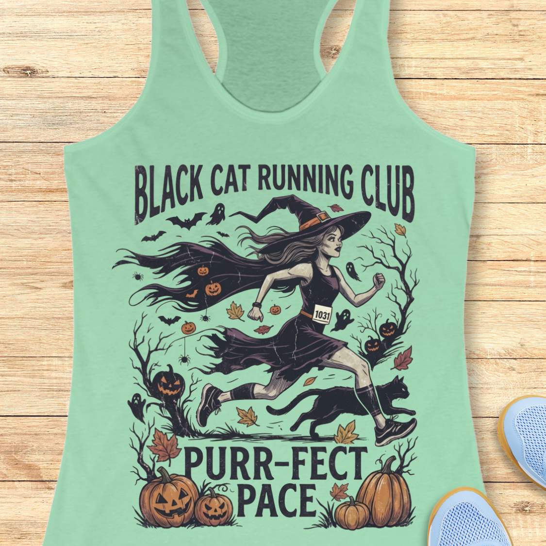 A mint green racerback tank top featuring a Halloween-themed graphic of a witch running with a black cat, pumpkins, bats, and the text 'BLACK CAT RUNNING CLUB' and 'PURR-FECT PACE'. The tank top is displayed flat on a light wood plank background with partial blue shoes visible in the corners.