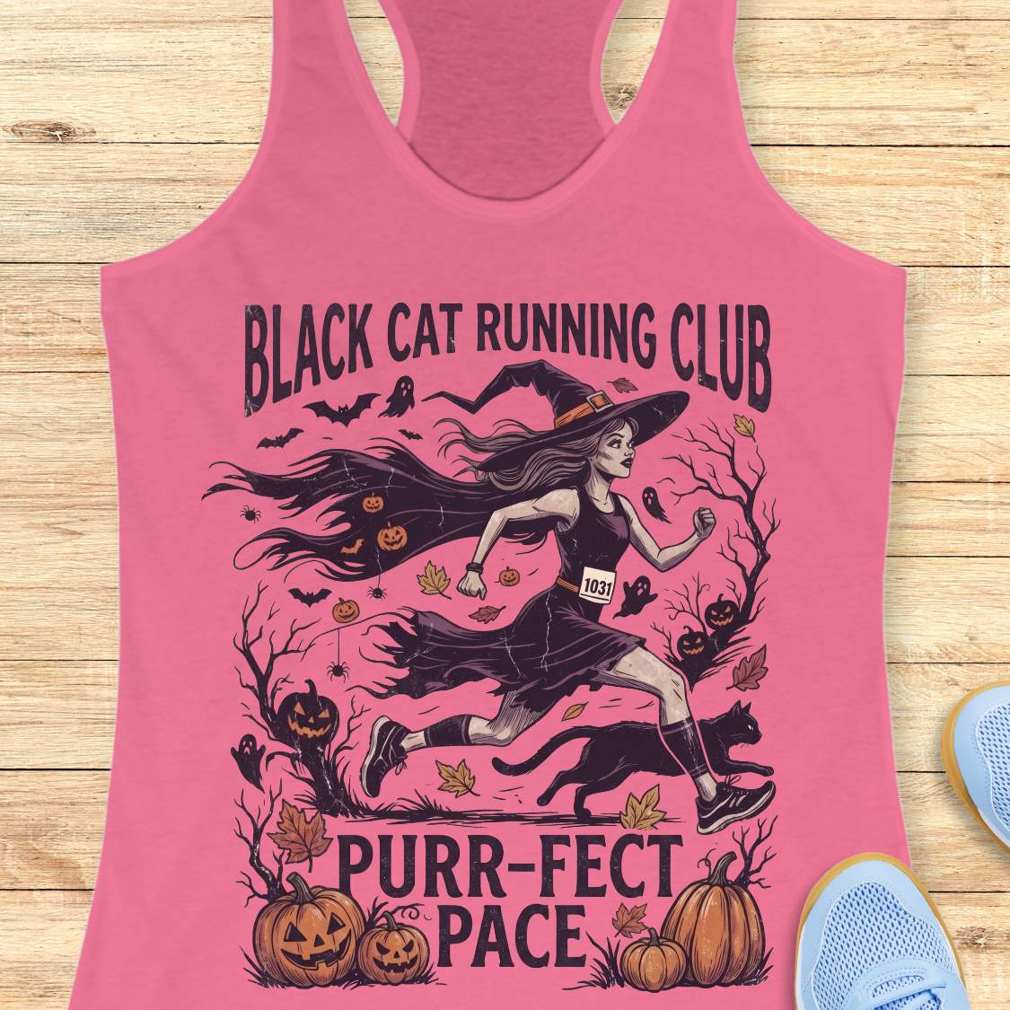 A pink racerback tank top laid flat on a light wood background, featuring a Halloween-themed graphic with the text 'BLACK CAT RUNNING CLUB' and 'PURR-FECT PACE.' The design shows a witch running with a black cat, pumpkins, bats, and autumn leaves.