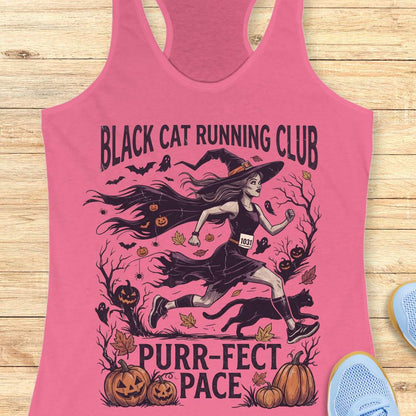 A pink racerback tank top laid flat on a light wood background, featuring a Halloween-themed graphic with the text 'BLACK CAT RUNNING CLUB' and 'PURR-FECT PACE.' The design shows a witch running with a black cat, pumpkins, bats, and autumn leaves.