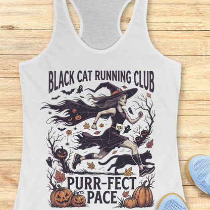 A white racerback tank top laid flat on a light wood background, featuring a Halloween-themed graphic with the text 'BLACK CAT RUNNING CLUB' and 'PURR-FACT PACE.' The design shows a witch running with a black cat, pumpkins, bats, leaves, and spooky trees.