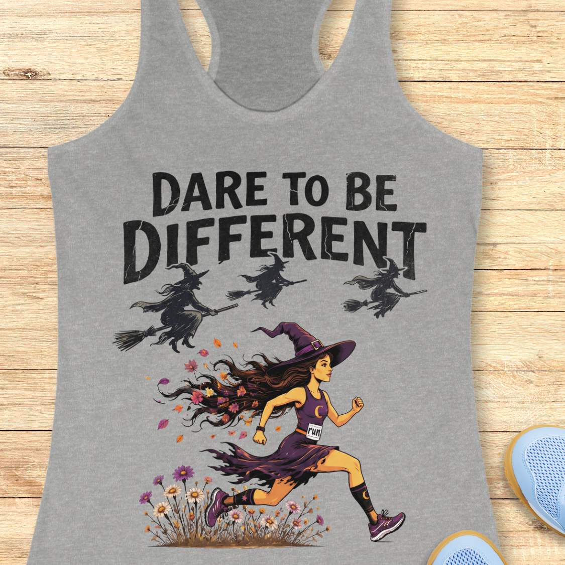 A heather gray racerback tank top with a graphic print of a witch running and three witches on broomsticks beneath the text 'DARE TO BE DIFFERENT'. The tank top is displayed flat on a light wood background with blue shoes partially visible in the corner.