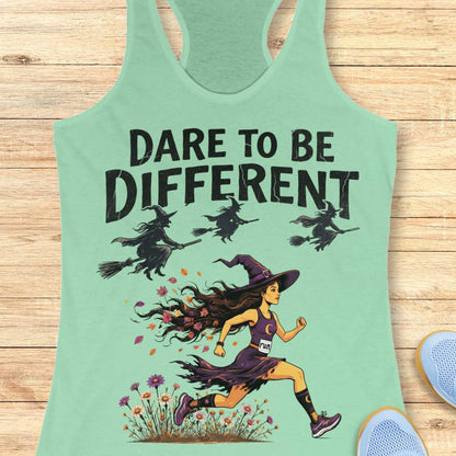 A mint green racerback tank top with a graphic print of a witch running through flowers with the text 'DARE TO BE DIFFERENT' and three witches flying on broomsticks above. The tank top is displayed flat on a light wood background with partial blue shoes visible in the corners.