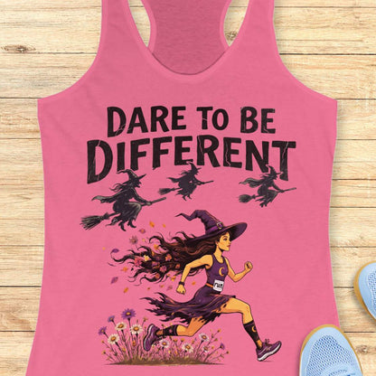 A pink racerback tank top laid flat on a light wood background, featuring a graphic of a witch running with broomsticks and the text 'DARE TO BE DIFFERENT'.
