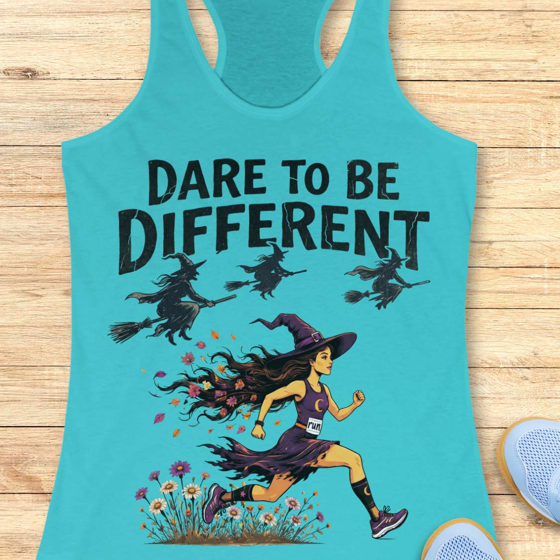A turquoise racerback tank top laid flat on a light wood surface, featuring a graphic print of a witch running through flowers with three witches on broomsticks and the text 'DARE TO BE DIFFERENT'.