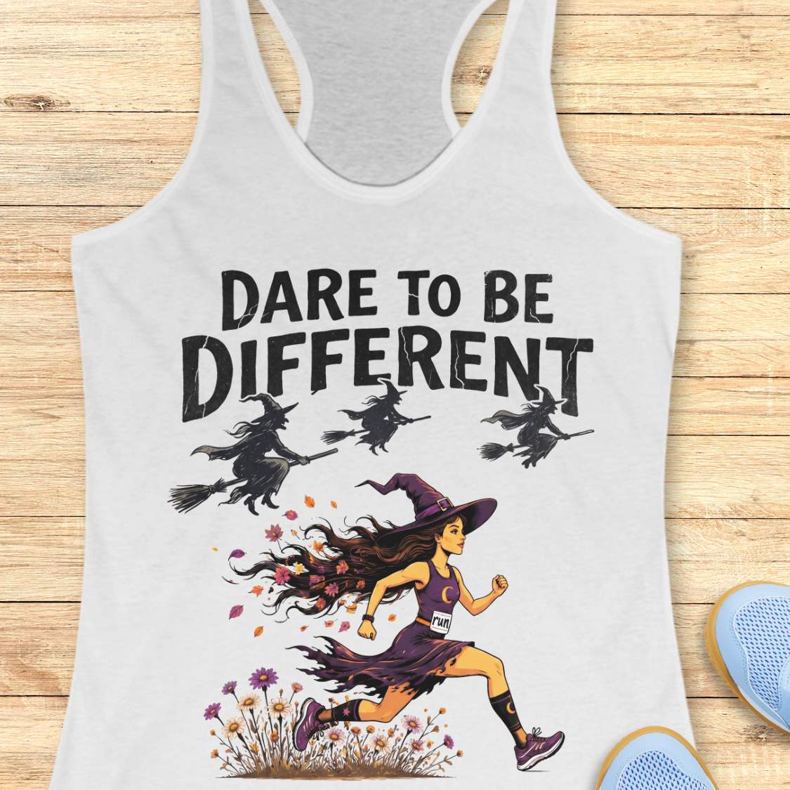 A white racerback tank top laid flat on a light wood background, featuring a graphic print of a witch running with the text 'DARE TO BE DIFFERENT' and three silhouettes of witches on broomsticks. Partial blue shoes are visible at the bottom right edge.