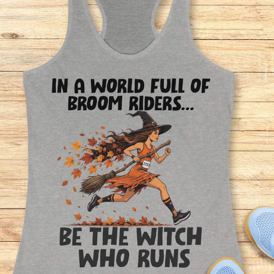 A heather gray racerback tank top featuring a graphic of a woman running on a broomstick with autumn leaves, with the text 'IN A WORLD FULL OF BROOM RIDERS.