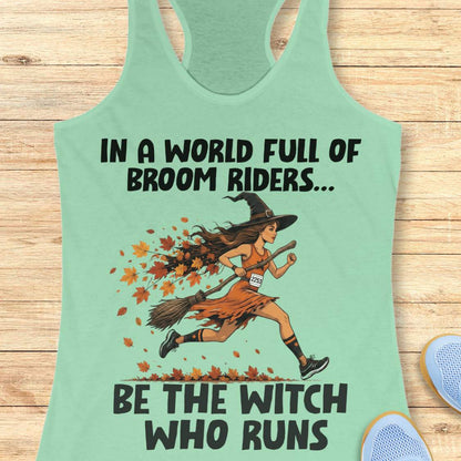 A mint green racerback tank top laid flat on a light wood background, featuring a graphic of a witch running on a broomstick with autumn leaves, accompanied by the text 'IN A WORLD FULL OF BROOM RIDERS.