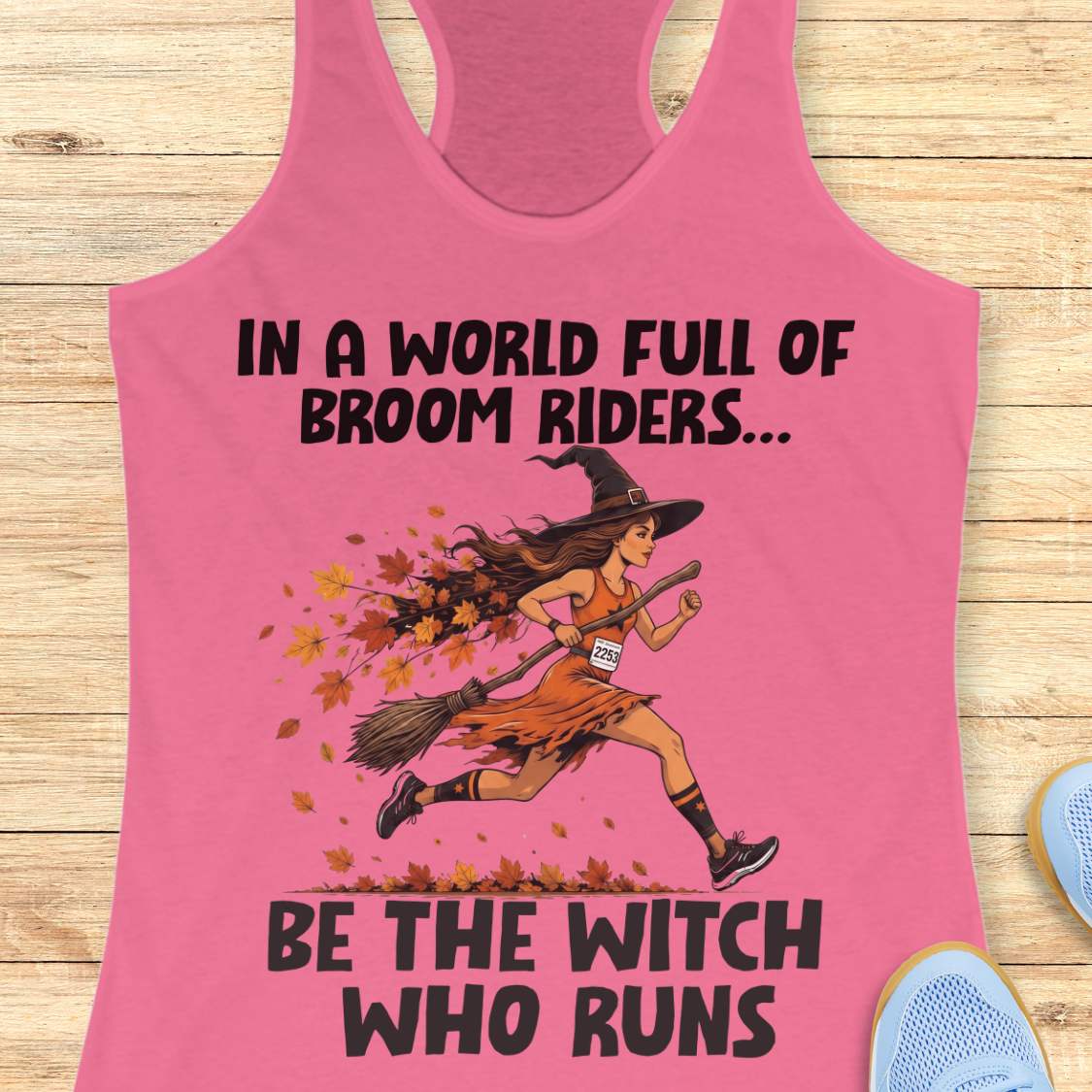A pink racerback tank top laid flat on a light wood background, featuring a graphic of a witch running with a broom and the text 'IN A WORLD FULL OF BROOM RIDERS.