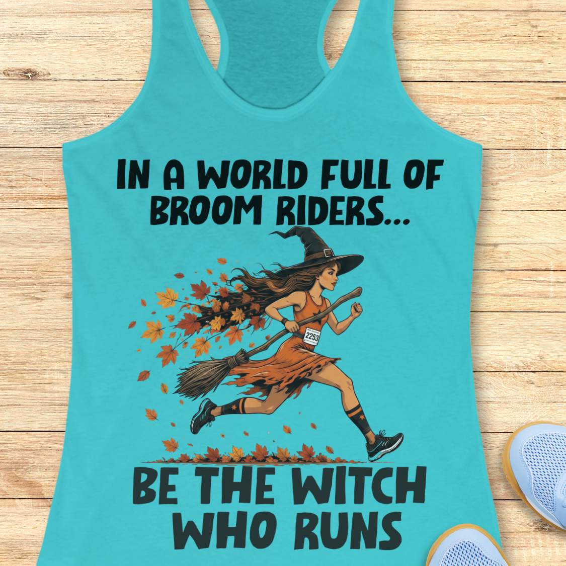 A turquoise racerback tank top laid flat on a light wood surface, featuring a printed graphic of a woman running as a witch with autumn leaves and the text 'IN A WORLD FULL OF BROOM RIDERS.
