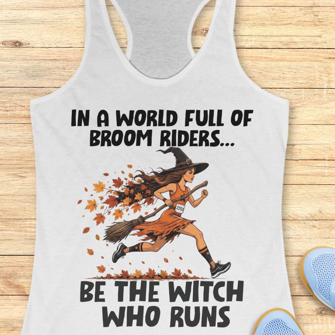 A white racerback tank top with a graphic print of a witch running on a broomstick with autumn leaves, accompanied by the text 'IN A WORLD FULL OF BROOM RIDERS.
