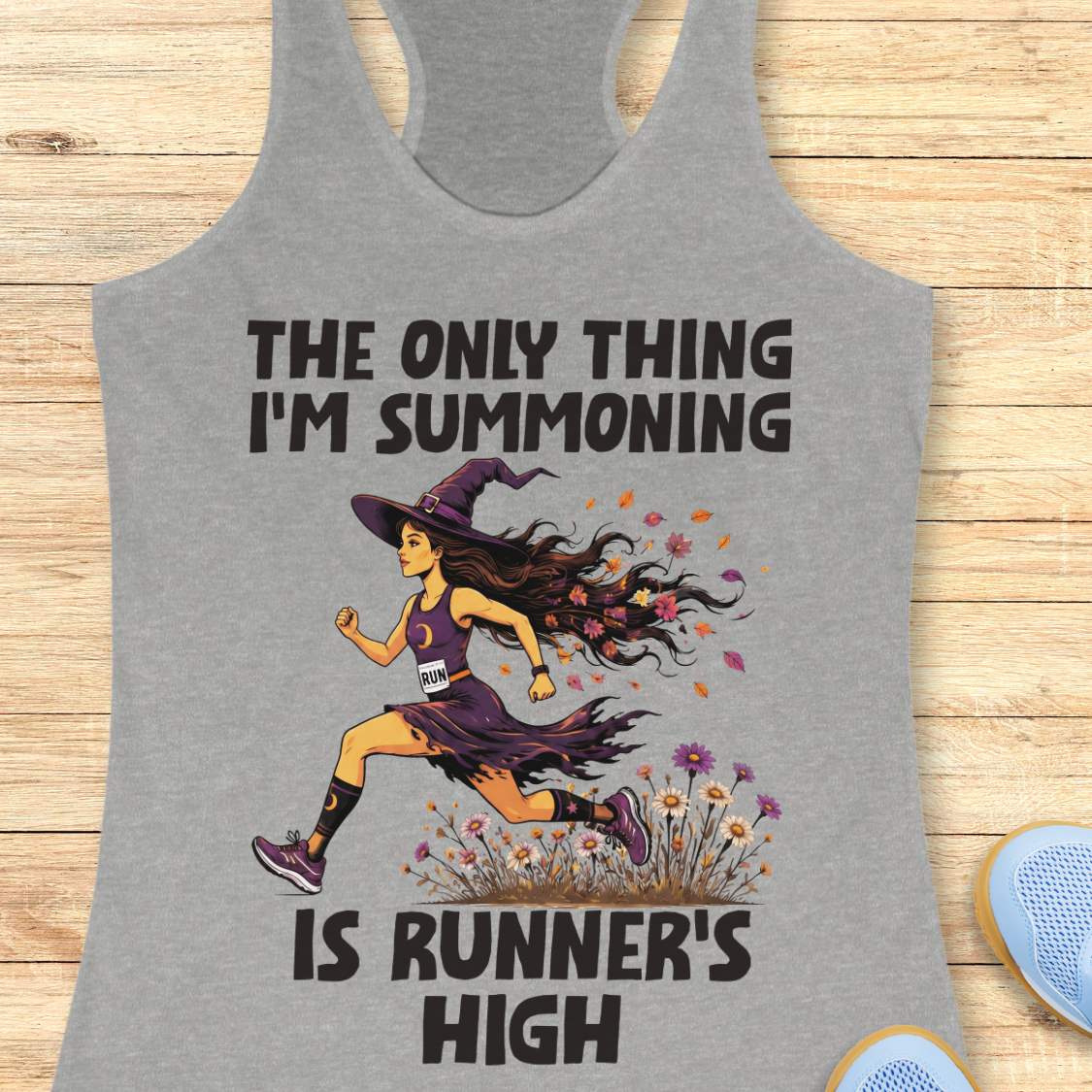A grey racerback tank top laid flat on a light wood surface, featuring a Halloween graphic of a witch running with a text 'The only thing I'm summoning is runner's high'.