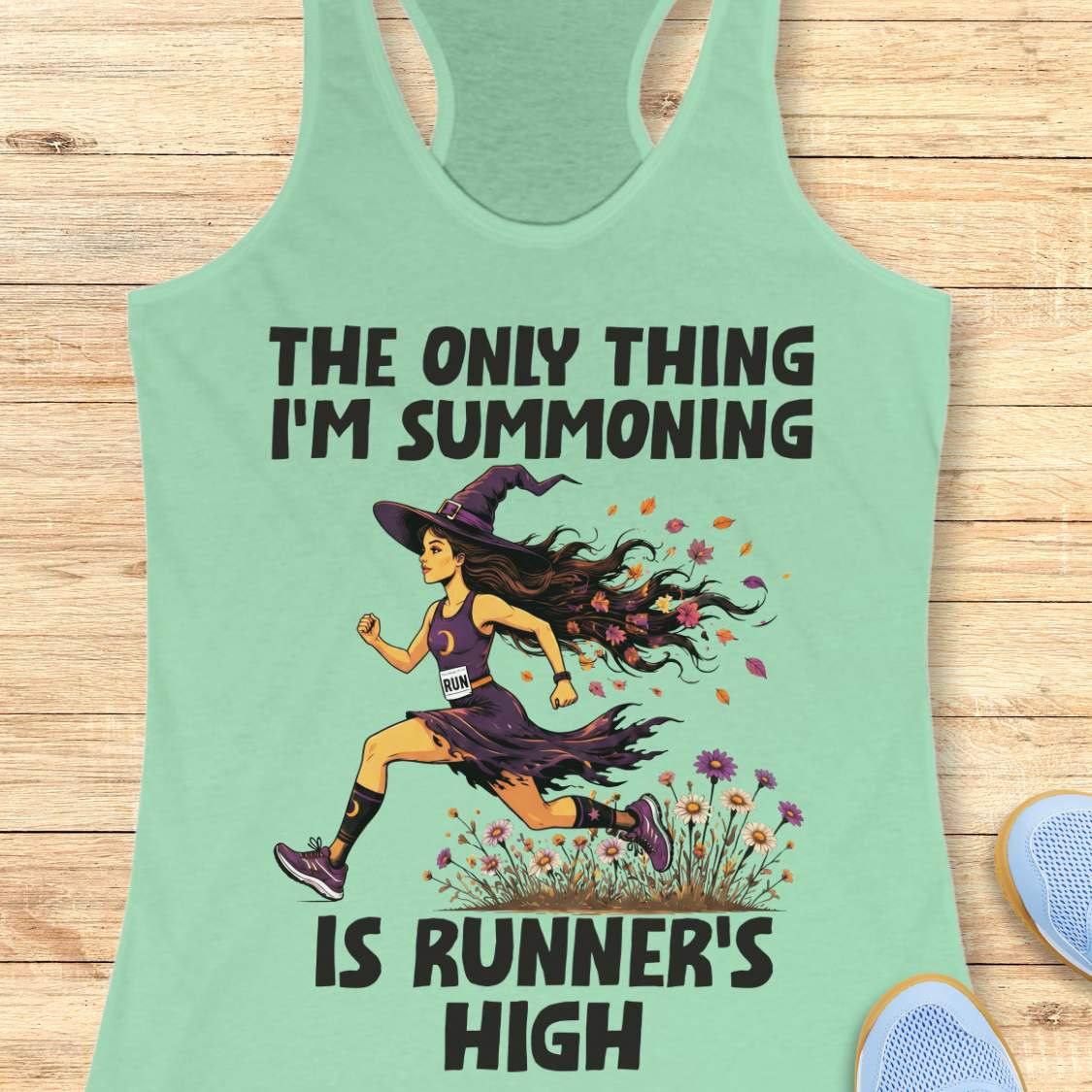 A mint racerback tank top laid flat on a light wood surface, featuring a Halloween graphic of a witch running with a text 'The only thing I'm summoning is runner's high'.