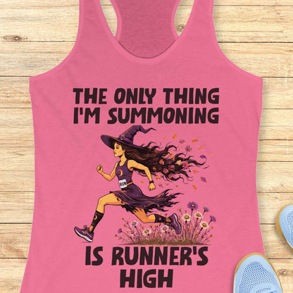 A pink racerback tank top laid flat on a light wood surface, featuring a Halloween graphic of a witch running with a text 'The only thing I'm summoning is runner's high'.