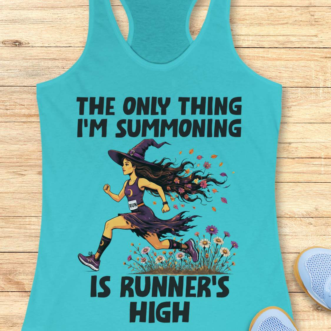 A blue racerback tank top laid flat on a light wood surface, featuring a Halloween graphic of a witch running with a text 'The only thing I'm summoning is runner's high'.