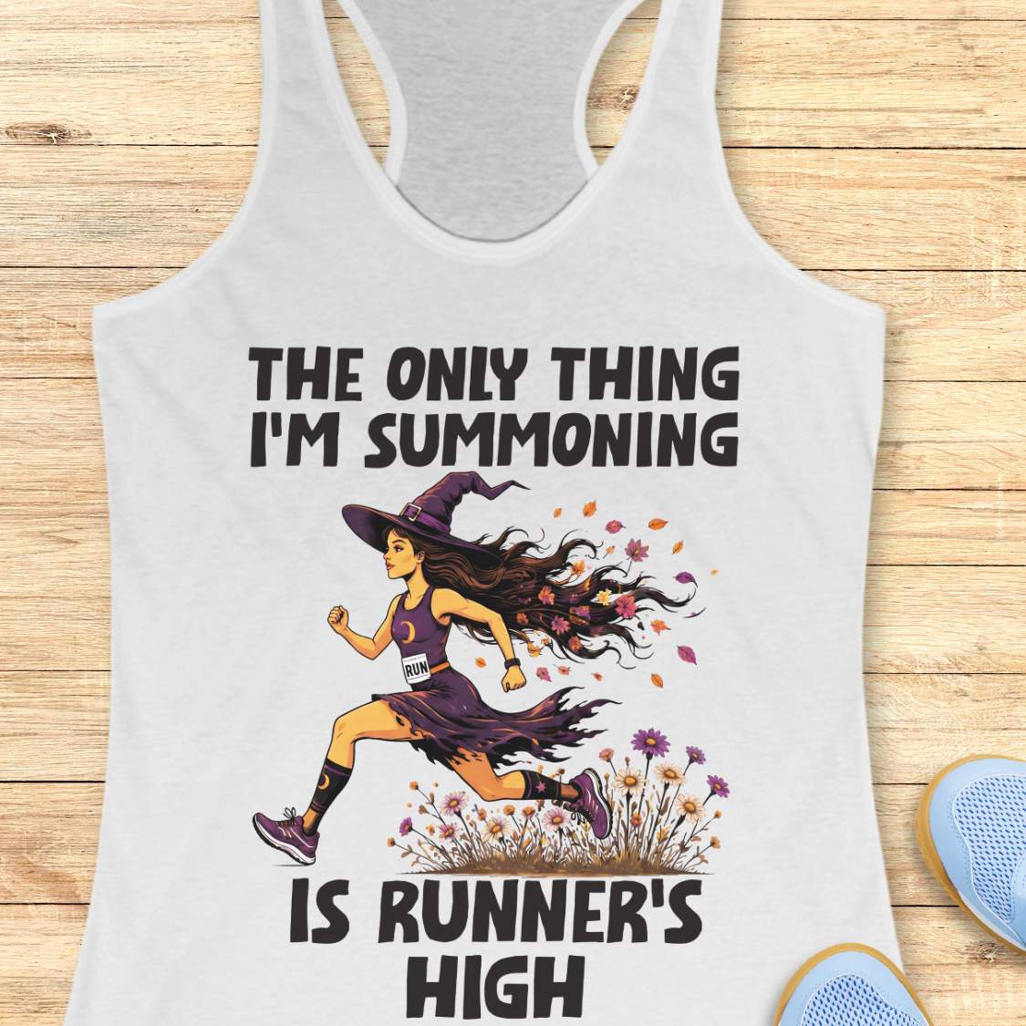 A white racerback tank top laid flat on a light wood surface, featuring a Halloween graphic of a witch running with a text 'The only thing I'm summoning is runner's high'.