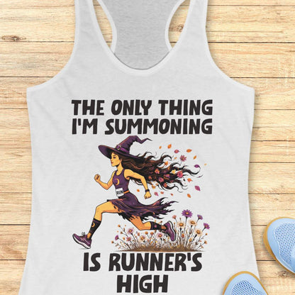 A white racerback tank top laid flat on a light wood surface, featuring a Halloween graphic of a witch running with a text 'The only thing I'm summoning is runner's high'.
