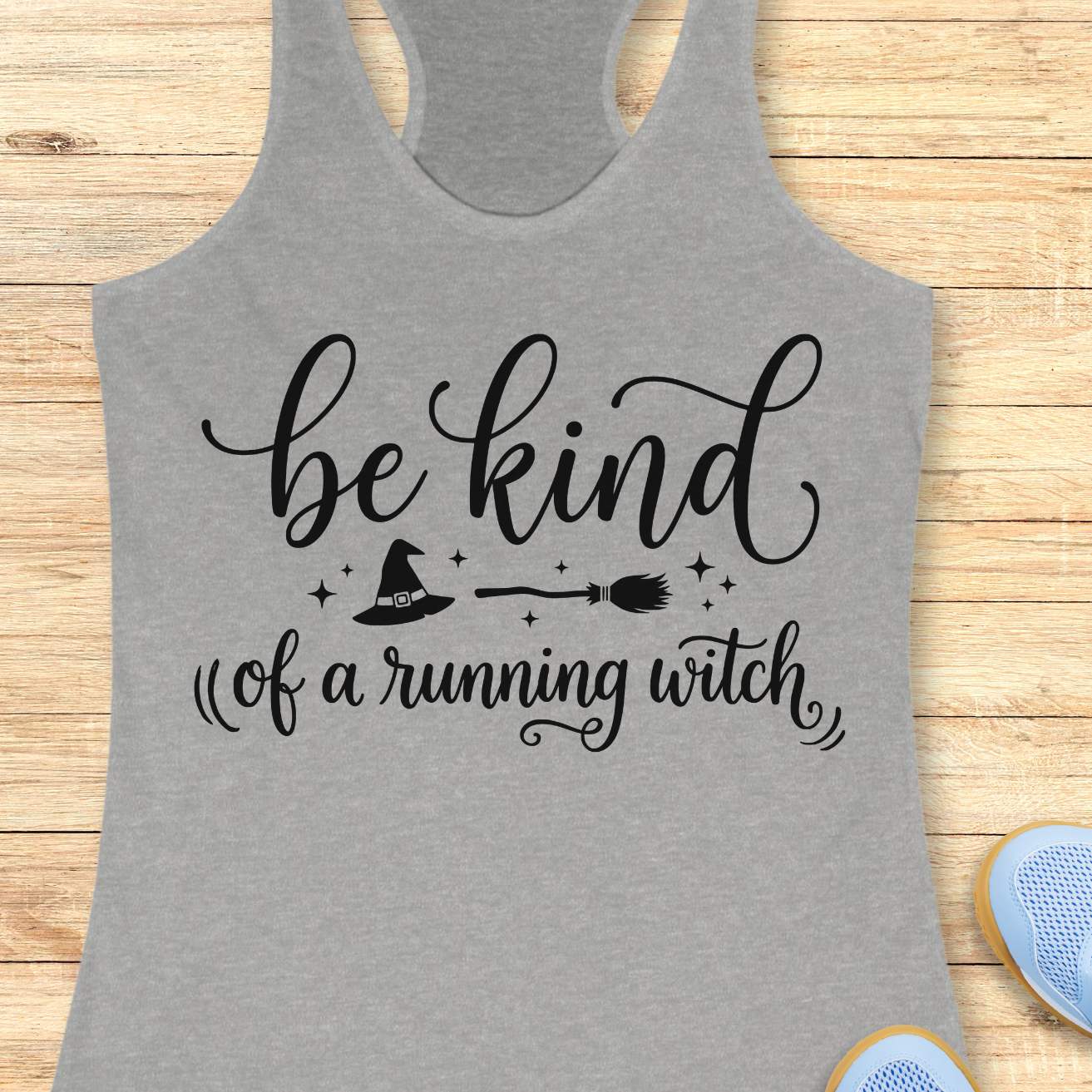 A heather gray racerback tank top with a black graphic print that reads 'be kind of a running witch' with a witch hat and broomstick illustration, laid flat on a light wood background.