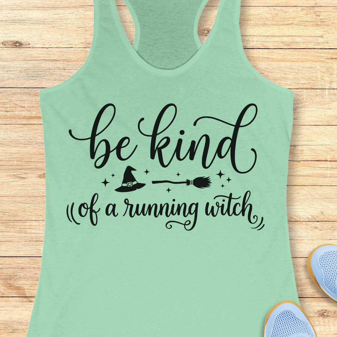 A mint green racerback tank top with a black graphic print reading 'be kind of a running witch' with a witch hat, broom, and stars, displayed flat on a light wood background with partial sneakers visible at the edges.