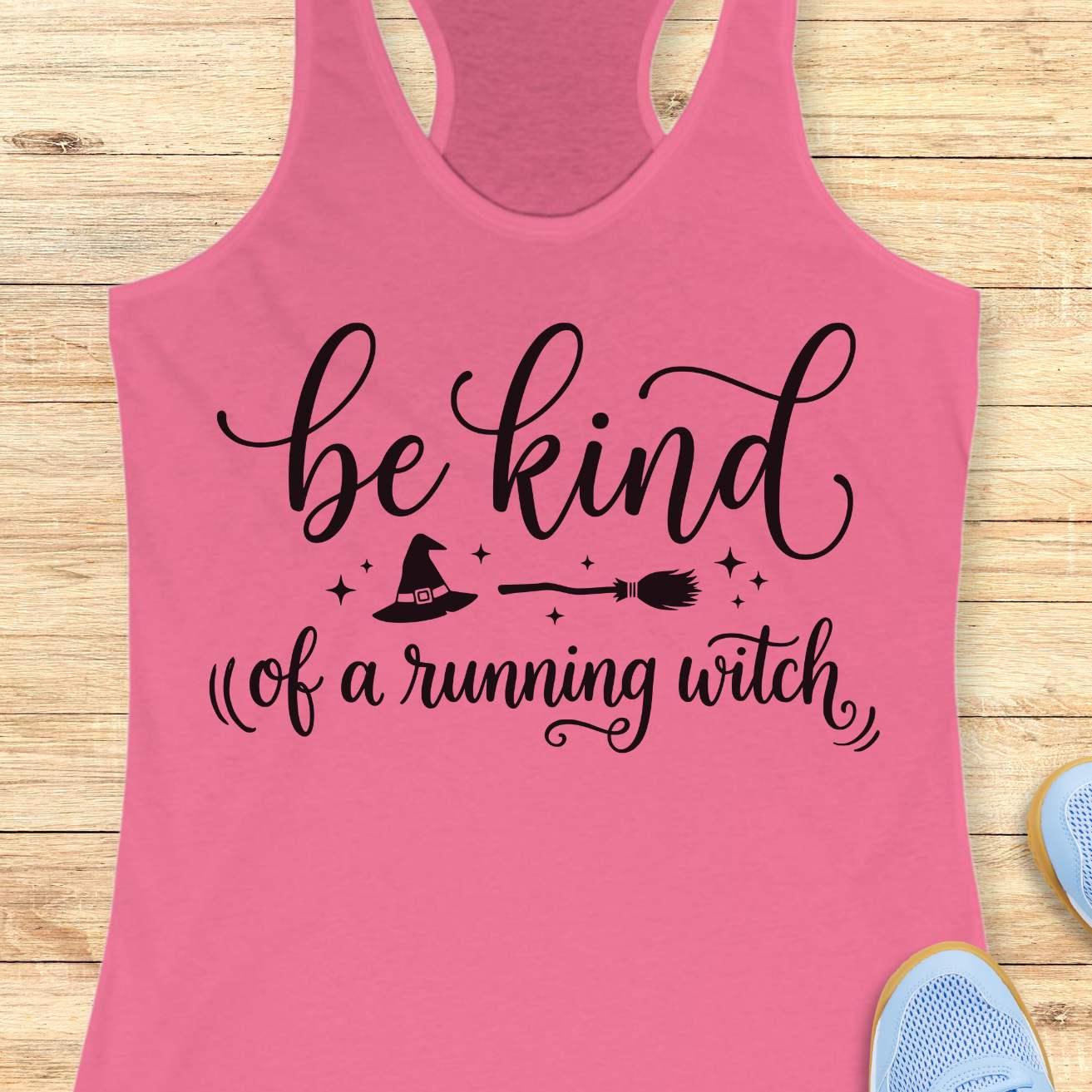A pink racerback tank top with a black graphic print reading 'be kind of a running witch' with a witch hat, broom, and stars, displayed flat on a light wood background with partial sneakers visible at the edges.