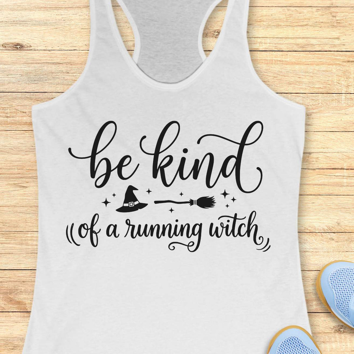 A white racerback tank top with a black graphic print that reads 'be kind of a running witch' with a witch hat, broom, and stars, displayed flat on a light wood background.