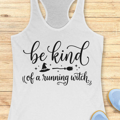A white racerback tank top with a black graphic print that reads 'be kind of a running witch' with a witch hat, broom, and stars, displayed flat on a light wood background.