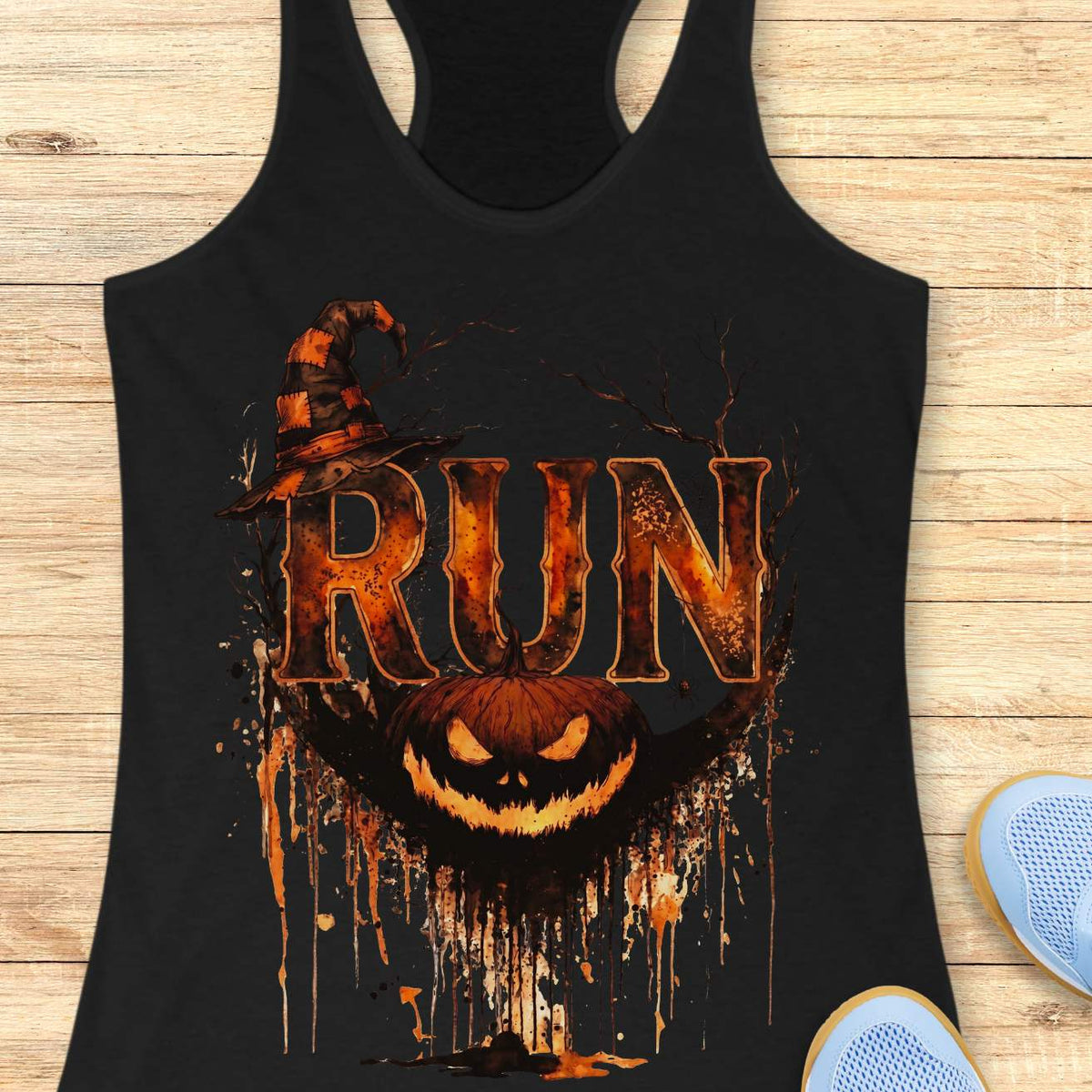 black racerback tank top featuring a Halloween-themed running design. The graphic displays the word “RUN” in large distressed orange and black letters, topped with a weathered witch hat. Behind the text are bare tree branches, a spider web, and a hanging spider. A grinning jack-o'-lantern with dripping black and orange paint effects sits below the lettering, creating a spooky autumn aesthetic. The tank top is displayed on a light wooden background with partial blue running shoes visible in the corners.