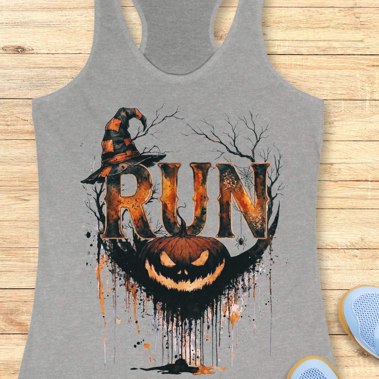 grey racerback tank top featuring a Halloween-themed running design. The graphic displays the word “RUN” in large distressed orange and black letters, topped with a weathered witch hat. Behind the text are bare tree branches, a spider web, and a hanging spider. A grinning jack-o'-lantern with dripping black and orange paint effects sits below the lettering, creating a spooky autumn aesthetic. The tank top is displayed on a light wooden background with partial blue running shoes visible in the corners.