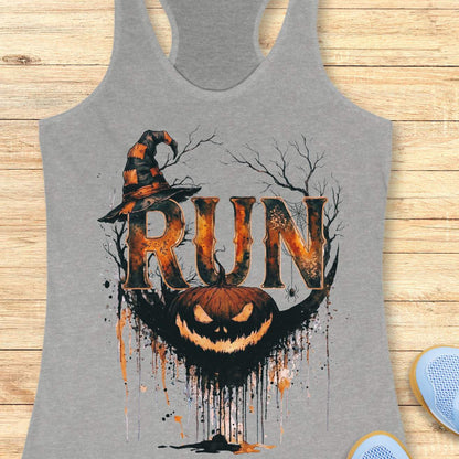 grey racerback tank top featuring a Halloween-themed running design. The graphic displays the word “RUN” in large distressed orange and black letters, topped with a weathered witch hat. Behind the text are bare tree branches, a spider web, and a hanging spider. A grinning jack-o'-lantern with dripping black and orange paint effects sits below the lettering, creating a spooky autumn aesthetic. The tank top is displayed on a light wooden background with partial blue running shoes visible in the corners.