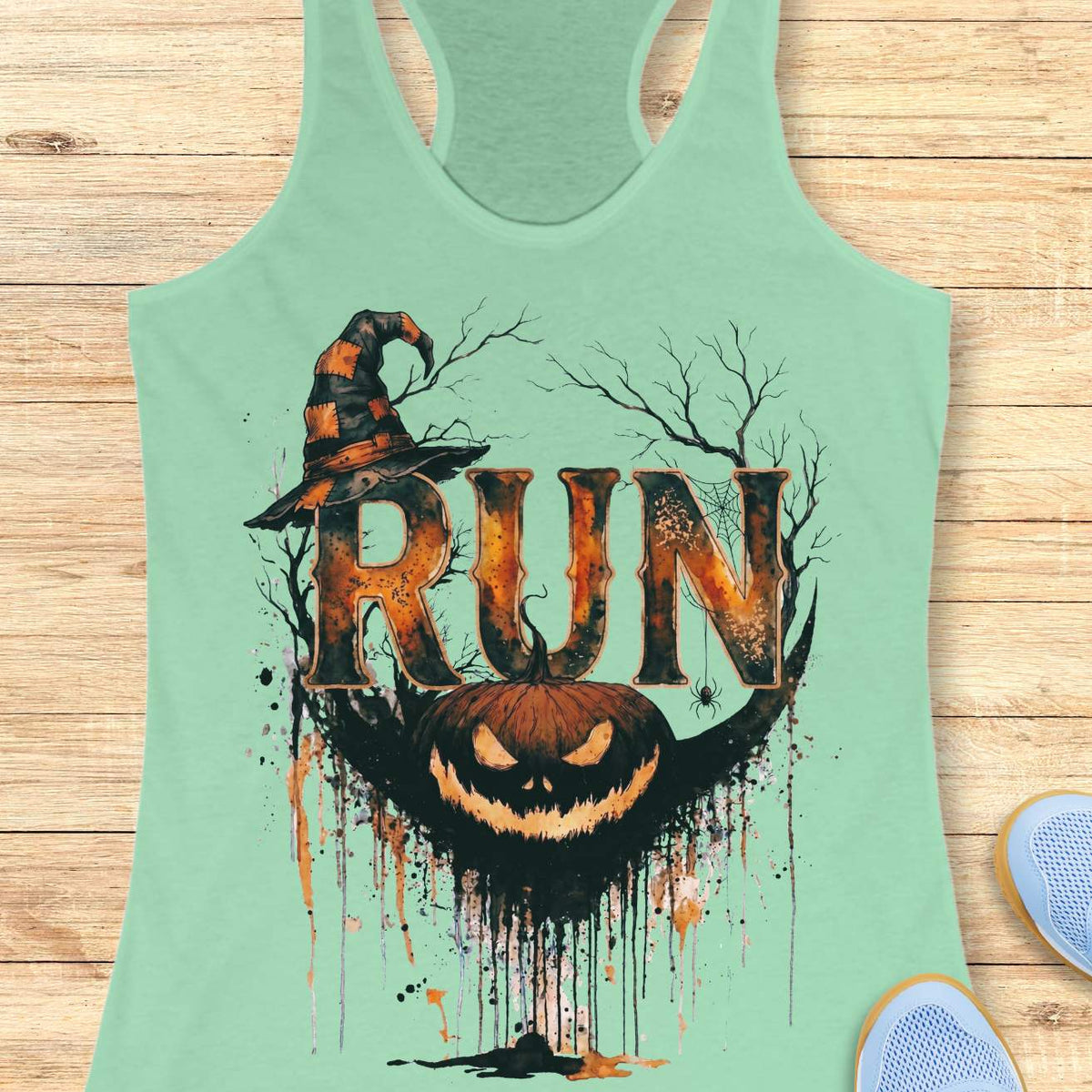 Mint green racerback tank top featuring a Halloween-themed running design. The graphic displays the word “RUN” in large distressed orange and black letters, topped with a weathered witch hat. Behind the text are bare tree branches, a spider web, and a hanging spider. A grinning jack-o'-lantern with dripping black and orange paint effects sits below the lettering, creating a spooky autumn aesthetic. The tank top is displayed on a light wooden background with partial blue running shoes visible in the corners.